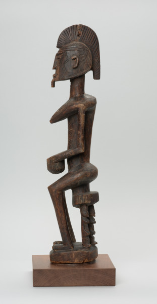 drewniana figura - Ujęcie lewego boku. Drewniana, rzeźbiona figura kobiety, siedzącej na stołku.