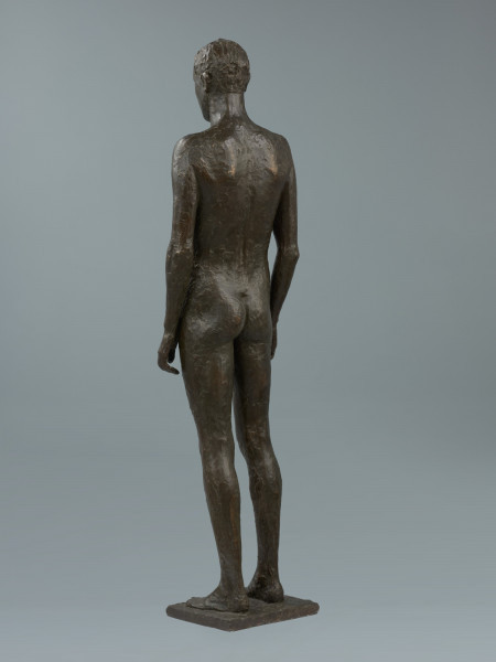 akt męski - Ujęcie z tyłu lekkim skosem w lewą stronę; Rzeźba z brązu przedstawiająca nagiego, młodego, szczupłego mężczyznę, stojącego twarzą do widza z rękami wzdłuż ciała.