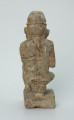 figura kultu przodków - Ujęcie z tyłu. Ustawiona na kwadratowej podstawie, wyrzeźbiona w biało-szarym kamieniu postać zamyślonego, siedzącego na stołku mężczyzny w czapce, o zaokrąglonych rogach i spłaszczonej części górnej.