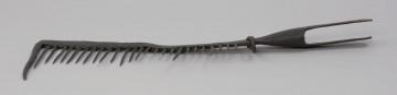 grzebień - Ujęcie z przodu. 26-zębna ość grzebieniasta, wykonana techniką kowalską. Ramie robocze płaskie, z jednego końca zagięte pod kątem 90 stopni (część ta ma formę zęba grubszego i dłuższego od pozostałych), w drugim końcu przechodzi w tuleję z dwoma pasami, służącą do osadzenia narzędzia na trzonku (trzonka brak). Na końcu każdego pasa otwór – jednego okrągły, drugiego – kwadratowy. Przy nasadzie tulei uszko wykonane z wygiętego w kształcie litery „U” pręta o okrągłym przekroju. Mocowane za pomocą nitu. Zęby mocowane w ramieniu, nierównej długości, ostro zakończone.  Ząb ósmy, licząc od uszka, przynitowany, długi.