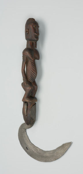 sierp - Ujęcie z przodu w pionie. Sierp o kutym z żelaza, łukowatym ostrzu z drewnianą rączką w formie rzeźbionej stojącej postaci z biustem i brodą z rękoma na udach.