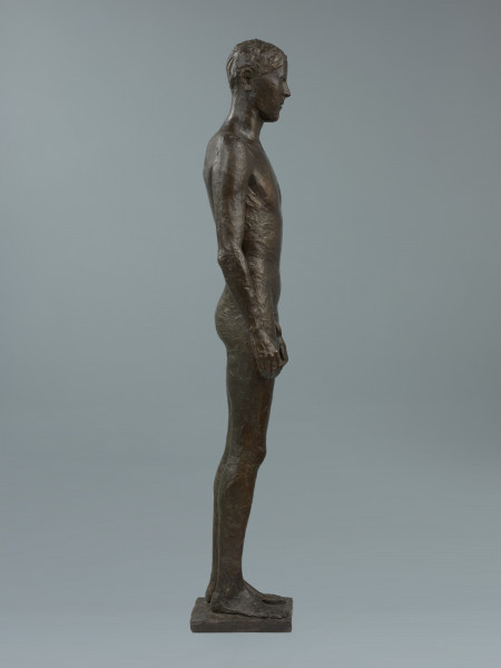 akt męski - Ujęcie z prawej strony; Rzeźba z brązu przedstawiająca nagiego, młodego, szczupłego mężczyznę, stojącego twarzą do widza z rękami wzdłuż ciała.