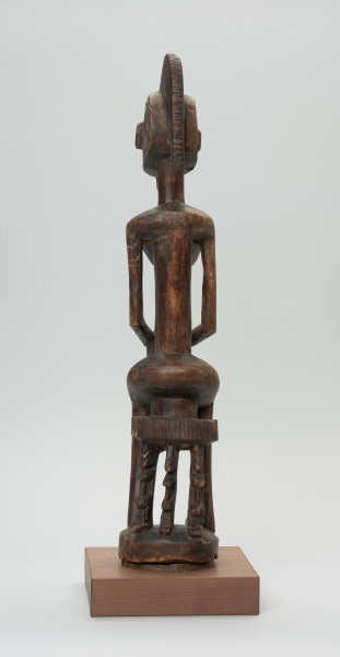 drewniana figura - Ujęcie z tyłu. Drewniana, rzeźbiona figura kobiety, siedzącej na stołku.