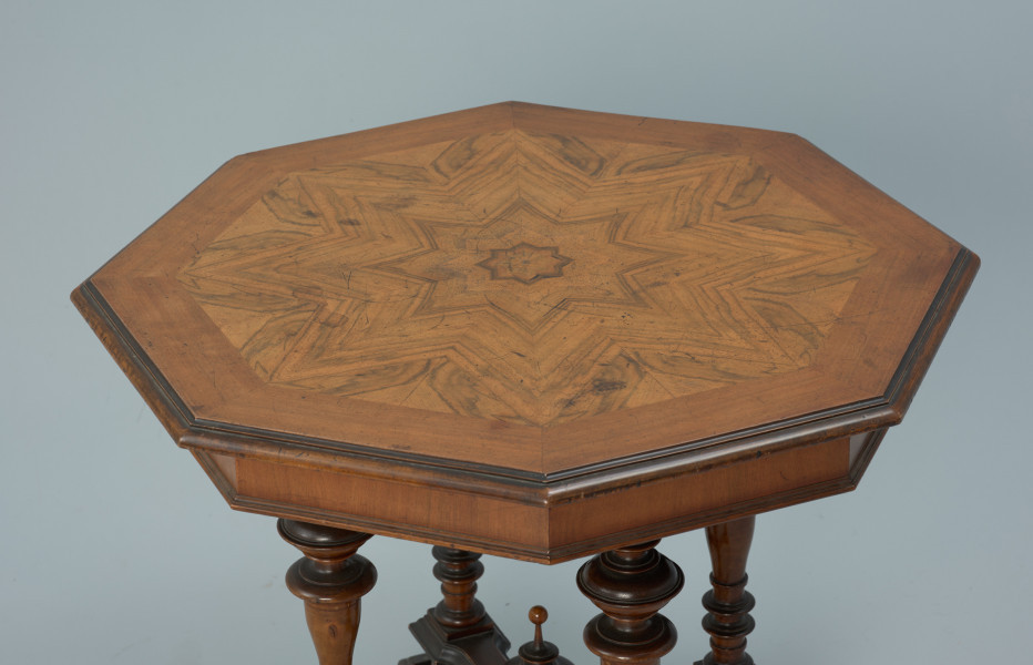Ośmiokątny, drewniany stolik - Ujęcie z góry, z ukosa. Stolik z ośmiokątnym blatem wykończonym fornirem układanym we wzór tworzący rozetę. Krawędzie sfazowane, profilowane. Skrzynia wspierająca blat ośmioboczna, wykończona profilowaną listwą.