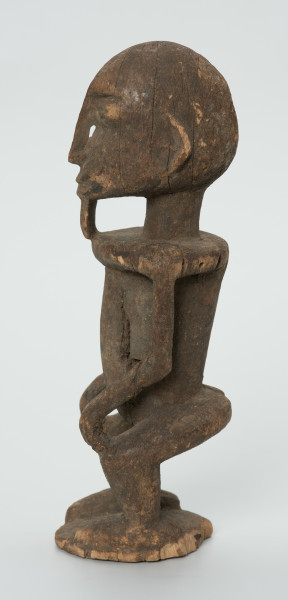 drewniana figura - Ujęcie prawego boku. Drewniana, rzeźbiona figura mężczyzny. Twarz owalna, długi nos i broda. Ręce oparte na kolanach. Ciało szczupłe.