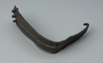 Fragment żelaznej klamry do pasa zdobiony żeberkami - Ujęcie z boku z lewej strony. Fragment żelaznej klamry do pasa zdobiony żeberkami. Od strony łącznika znajdują się trzy nity.