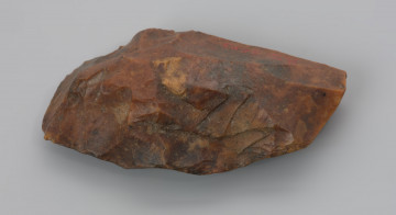 Mały ciosak rdzeniowy - ujęcie z boku; Na zdjęciu ciosak rdzeniowy, narzędzie wykonano z krzemienia kredowego. Pokryte jest czerwonopomarańczową patyną, która jest efektem zalegania w środowisku bogatym w związki żelaza.