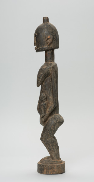 rzeźba - Ujęcie lewego boku. Drewniana, rzeźbiona postać kobiety w pozycji stojącej z dzieckiem ułożonym na lewym biodrze. Głowa stosunkowo duża, tułów zaokrąglony. Oczy, nos (w kształcie strzały), oczy broda lekko zarysowane.