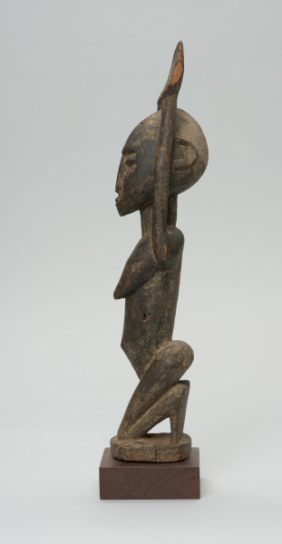 rzeźbiona figura - Ujęcie lewego boku. Drewniana, rzeźbiona postać kobiety, znajdującej się w pozycji siedzącej. Jej obie ręce skierowane są do góry.