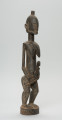 rzeźba - Ujęcie z przodu z prawej strony. Drewniana, rzeźbiona postać kobiety w pozycji stojącej z dzieckiem ułożonym na lewym biodrze. Głowa stosunkowo duża, tułów zaokrąglony. Oczy, nos (w kształcie strzały), oczy broda lekko zarysowane.