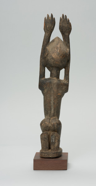 rzeźbiona figura - Ujęcie z tyłu. Drewniana, rzeźbiona postać kobiety, znajdującej się w pozycji siedzącej. Jej obie ręce skierowane są do góry.