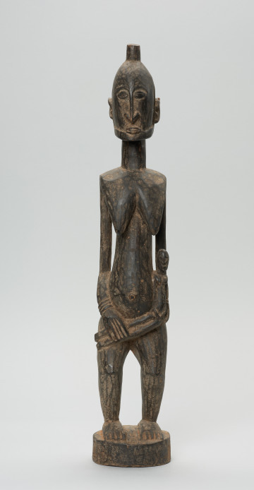 rzeźba - Ujęcie z przodu. Drewniana, rzeźbiona postać kobiety w pozycji stojącej z dzieckiem ułożonym na lewym biodrze. Głowa stosunkowo duża, tułów zaokrąglony. Oczy, nos (w kształcie strzały), oczy broda lekko zarysowane.