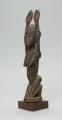 rzeźbiona figura - Ujęcie z przodu, z lewej strony. Drewniana, rzeźbiona postać kobiety, znajdującej się w pozycji siedzącej. Jej obie ręce skierowane są do góry.