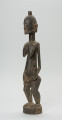 rzeźba - Ujęcie z przodu z lewej strony. Drewniana, rzeźbiona postać kobiety w pozycji stojącej z dzieckiem ułożonym na lewym biodrze. Głowa stosunkowo duża, tułów zaokrąglony. Oczy, nos (w kształcie strzały), oczy broda lekko zarysowane.