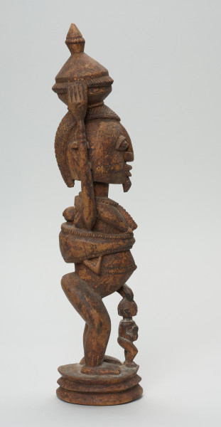 rzeźbiona figura - Ujęcie prawego boku. Drewniana, rzeźbiona postać kobiety stojącej z naczyniem na głowie oraz dziećmi. Jedno z dzieci na plecach, drugie (nieproporcjonalnie małe) stoi tuż przed kobietą.