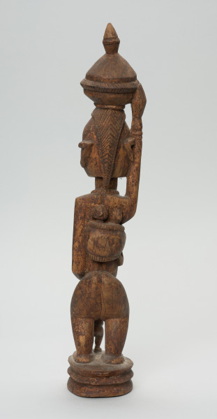rzeźbiona figura - Ujęcie z tyłu. Drewniana, rzeźbiona postać kobiety stojącej z naczyniem na głowie oraz dziećmi. Jedno z dzieci na plecach, drugie (nieproporcjonalnie małe) stoi tuż przed kobietą.