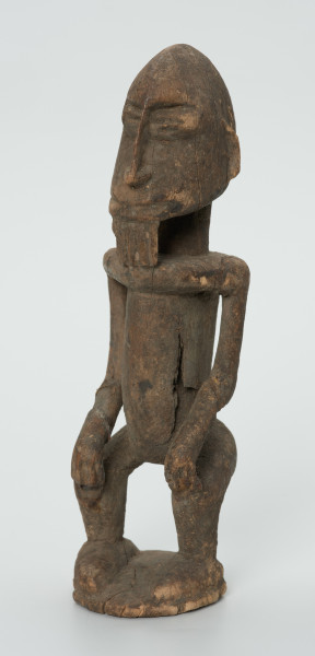 drewniana figura - Ujęcie z przodu skosem w lewą stronę. Drewniana, rzeźbiona figura mężczyzny. Twarz owalna, długi nos i broda. Ręce oparte na kolanach. Ciało szczupłe.