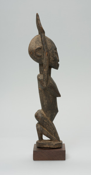 rzeźbiona figura - Ujęcie prawego boku. Drewniana, rzeźbiona postać kobiety, znajdującej się w pozycji siedzącej. Jej obie ręce skierowane są do góry.