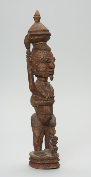 rzeźbiona figura - Ujęcie z przodu, z prawej strony. Drewniana, rzeźbiona postać kobiety stojącej z naczyniem na głowie oraz dziećmi. Jedno z dzieci na plecach, drugie (nieproporcjonalnie małe) stoi tuż przed kobietą.