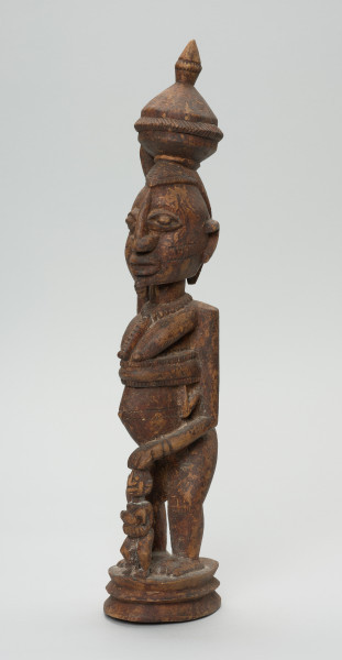 rzeźbiona figura - Ujęcie z przodu, z lewej strony. Drewniana, rzeźbiona postać kobiety stojącej z naczyniem na głowie oraz dziećmi. Jedno z dzieci na plecach, drugie (nieproporcjonalnie małe) stoi tuż przed kobietą.