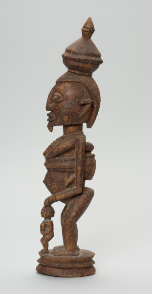 rzeźbiona figura - Ujęcie lewego boku. Drewniana, rzeźbiona postać kobiety stojącej z naczyniem na głowie oraz dziećmi. Jedno z dzieci na plecach, drugie (nieproporcjonalnie małe) stoi tuż przed kobietą.