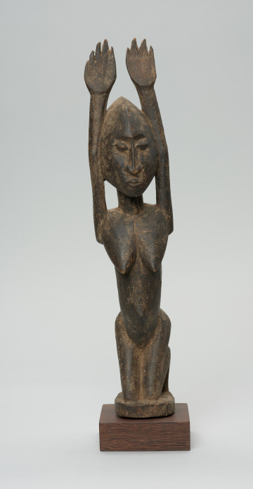 rzeźbiona figura - Ujęcie z przodu. Drewniana, rzeźbiona postać kobiety, znajdującej się w pozycji siedzącej. Jej obie ręce skierowane są do góry.