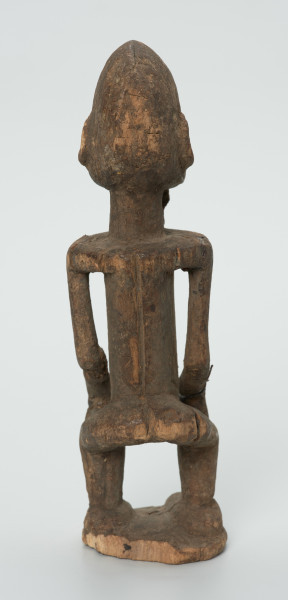 drewniana figura - Ujęcie z tyłu. Drewniana, rzeźbiona figura mężczyzny. Twarz owalna, długi nos i broda. Ręce oparte na kolanach. Ciało szczupłe.