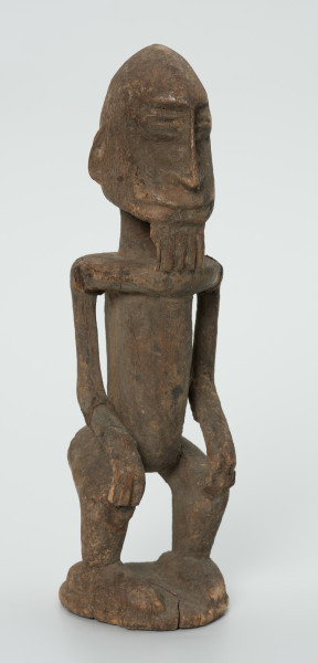 drewniana figura - Ujęcie z przodu skosem w prawą stronę. Drewniana, rzeźbiona figura mężczyzny. Twarz owalna, długi nos i broda. Ręce oparte na kolanach. Ciało szczupłe.