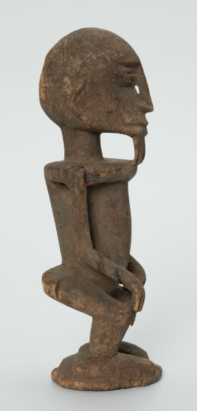 drewniana figura - Ujęcie lewego boku. Drewniana, rzeźbiona figura mężczyzny. Twarz owalna, długi nos i broda. Ręce oparte na kolanach. Ciało szczupłe.