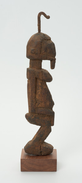 rzeźba - Ujęcie z leej strony. Drewniana rzeźba postaci męskiej. Wąska podłużna twarz, na głowie haczykowata ozdoba. Tułów długi, nogi zgięte w kolanach, ręce oparte na kolanach.