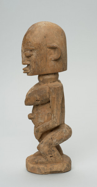 rzeźba - Ujęcie z lewego boku; Figura - postać kobiety w pozycji stojącej. Sylwetka masywna, przysadzista. Głowa duża, z wyrzeźbionymi uszami. Ręce i piersi wydobyte z bryły głębokimi nacięciami. Dłonie ułożone na łonie. Widoczne stopy. Związana z kultem przodków.