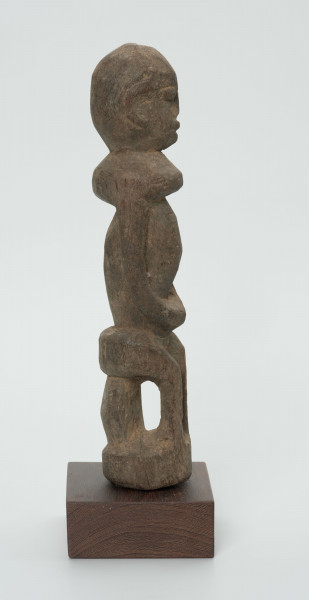 Drewniana figurka postaci siedzącej na stołku - Ujęcie z prawego boku; postać siedząca na stołku. Części ciała i twarz schematyczne, zaznaczone płytkimi nacięciami. Figura statyczna.