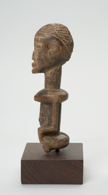 rzeźba - Ujęcie lewego boku. Figura przedstawiająca postać kobiety. Duża głowa, z zaznaczoną fryzurą, ręce prostymi cięciami wydobyte z tułowia. Figura kanoniczna. Brak nóg. Związana z kultem przodków.