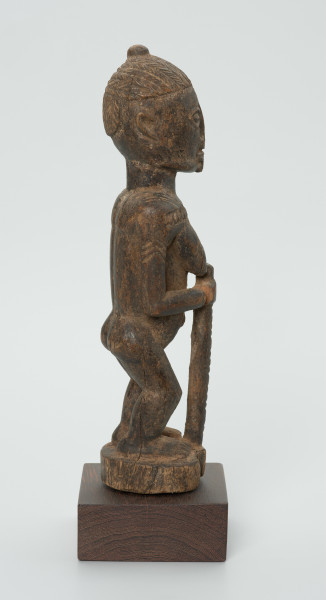 Drewniana figurka kobiety z laską - Ujęcie z prawego boku. Drewniana figurka kobiety w pozycji stojącej. W prawej dłoni trzyma laskę. Uszkodzona lewa dłoń. Całość pokryta drobnymi wzorami łącznie z włosami upiętymi w kok.
