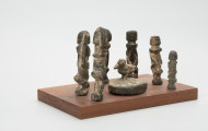 rzeźba - Ujęcie z lewego boku; Zestaw składa się z ośmiu małych, drewnianych figurek w pozycji stojącej: siedmiu antropomorficznych i jednej zoomorficznej - przedstawiającej ptaka (dzioborożca calao) na dużym, zaokrąglonym cokoliku. Figurki postaci ludzkich wykonano w sposób schematyczny, dwie mają uniesione do góry ręce. Rzeźby pokryto dość sporą warstwą białej substancji ofiarnej tak grubej, że przesłania ich rzeźbiarską formę.