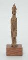 Drewniana figurka siedzącej postaci - Ujęcie z przodu; figurka przedstawiająca postać ludzką w pozycji siedzącej. Części ciała i rysy twarzy wydobyte za pomocą płytkich nacięć siekierki.