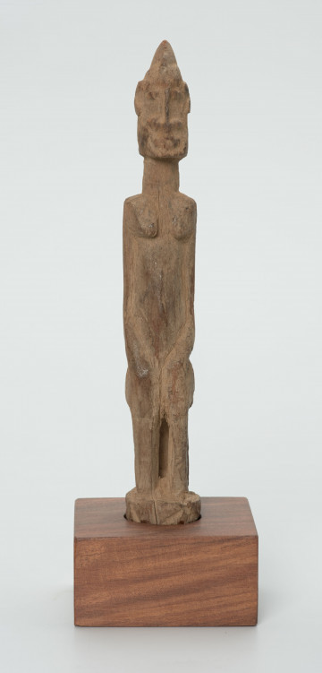 Drewniana figurka siedzącej postaci - Ujęcie z przodu; figurka przedstawiająca postać ludzką w pozycji siedzącej. Części ciała i rysy twarzy wydobyte za pomocą płytkich nacięć siekierki.