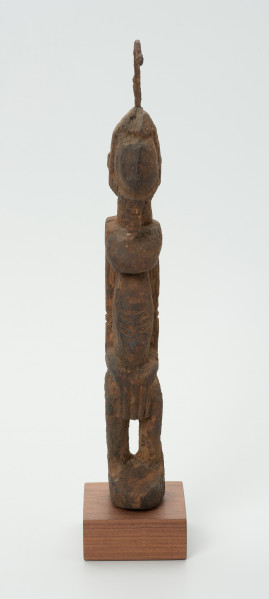 rzeźba - Ujęcie z przodu. Drewniana rzeźba postaci męskiej. Wąska podłużna twarz, na głowie haczykowata ozdoba. Tułów długi, nogi zgięte w kolanach, ręce oparte na kolanach.
