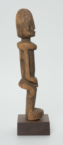 Drewniana figurka kobiety ze spiczastą głową - Ujęcie z prawego boku. Drewniana figurka kobiety w pozycji stojącej. Głowa duża, szpiczasta. Twarz, piersi, ręce schematyczne, wydobyte z bryły drewna płytkimi cięciami. Nogi w formie prostokątnej, lekko wygięte.