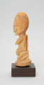 rzeźba - Ujęcie lewego boku. Figurka przedstawiająca kobietę siedzącą na podkulonych nogach. Głowa duża, reszta postaci wydobyta płytkimi nacięciami. Forma zgeometryzowana.