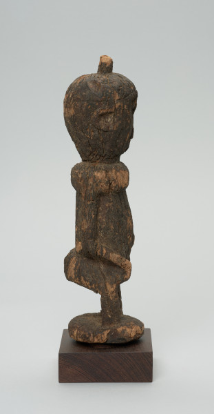 rzeźba - Ujęcie z lewej strony. Figurka - postać kobiety w pozycji stojącej. Głowa duża wychodząca bezpośrednio z tułowia. Ręce przylegające do tułowia. Na głowie mała wypustka.