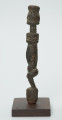 rzeźba - Ujęcie prawej strony rzeźby. Drewniana rzeźba postaci mężczyzny ze zgietymi kolanami. Schematyczne zarysowanie dużej głowy, długiej szyi, na której widoczny jest szeroki naszyjnik. Wąski, długi tułów, długie ręce oparte na kolanach, krótkie nogi i szerokie stopy.
