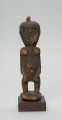rzeźba - Ujęcie z przodu. Figurka - postać kobiety w pozycji stojącej. Głowa duża wychodząca bezpośrednio z tułowia. Ręce przylegające do tułowia. Na głowie mała wypustka.