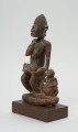 rzeźba - Ujęcie ze skosu z prawej;  Figura - kobieta z dziećmi. Rzeźba przedstawia siedzącą kobietę z niemowlakiem na rękach, które opiera na kolanach. Przy niej siedzi drugie dziecko, które kuca. Postać kobiety miejscami rzeźbiona symbolem zygzaka.