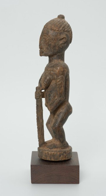 Drewniana figurka kobiety z laską - Ujęcie z lewego boku. Drewniana figurka kobiety w pozycji stojącej. W prawej dłoni trzyma laskę. Uszkodzona lewa dłoń. Całość pokryta drobnymi wzorami łącznie z włosami upiętymi w kok.