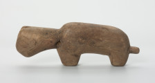 Drewniana figura zwierzęcia z krótkim ogonem - Ujęcie z lewej strony. Drewniana figurka nieokreślonego zwierzęcia z długą szyją, podłużną głową i krótkim ogonem.