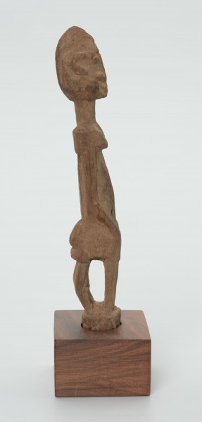 Drewniana figurka siedzącej postaci - Ujęcie z prawego boku; figurka przedstawiająca postać ludzką w pozycji siedzącej. Części ciała i rysy twarzy wydobyte za pomocą płytkich nacięć siekierki.