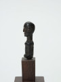 Mała drewniana figurka przedstawiająca popiersie człowieka - Ujęcie z lewej strony; Mała drewniana figurka przedstawiająca popiersie człowieka. Głowa i tułów starannie wygładzone. Postać ujęta schematycznie. Głowa, rysy, piersi, ręce, wydobyte płytkimi nacięciami.