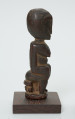 rzeźba - Ujęcie lewej strony rzeźby. Figurka - postać kobiety siedzącej na stołku. O jej płci informują wyraźnie zaznaczone piersi.