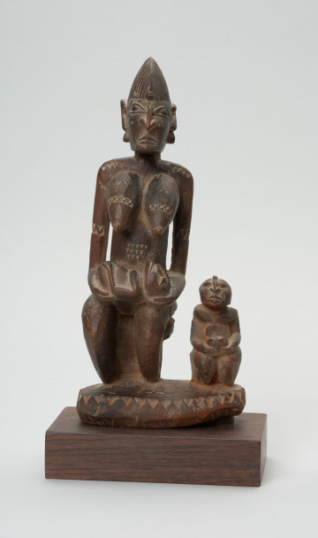 rzeźba - Ujęcie z przodu. Figura - kobieta z dziećmi. Rzeźba przedstawia siedzącą kobietę z niemowlakiem na rękach, które opiera na kolanach. Przy niej siedzi drugie dziecko, które kuca. Postać kobiety miejscami rzeźbiona symbolem zygzaka.